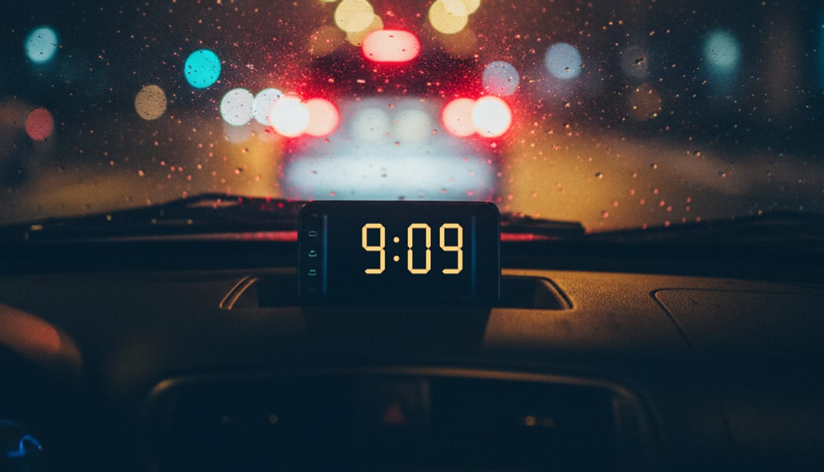A car dashboard clock reading 9:09 at night, seen from the driver's seat behind a rain-speckled windshield.
